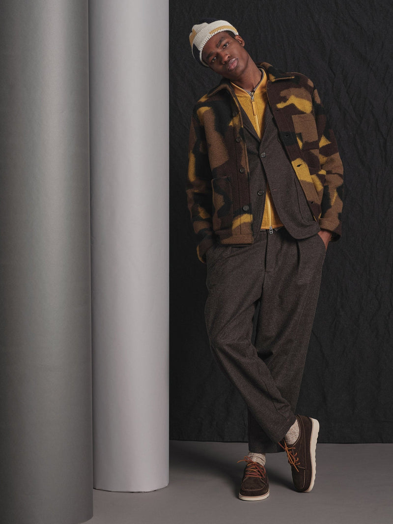 Man wearing a camouflage jacket and brown pants leaning against a gray column.