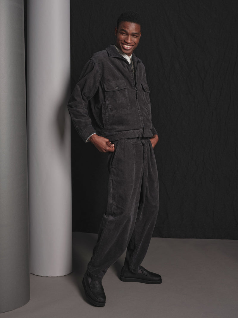 Man wearing a dark gray corduroy matching jacket and trousers standing against a black background