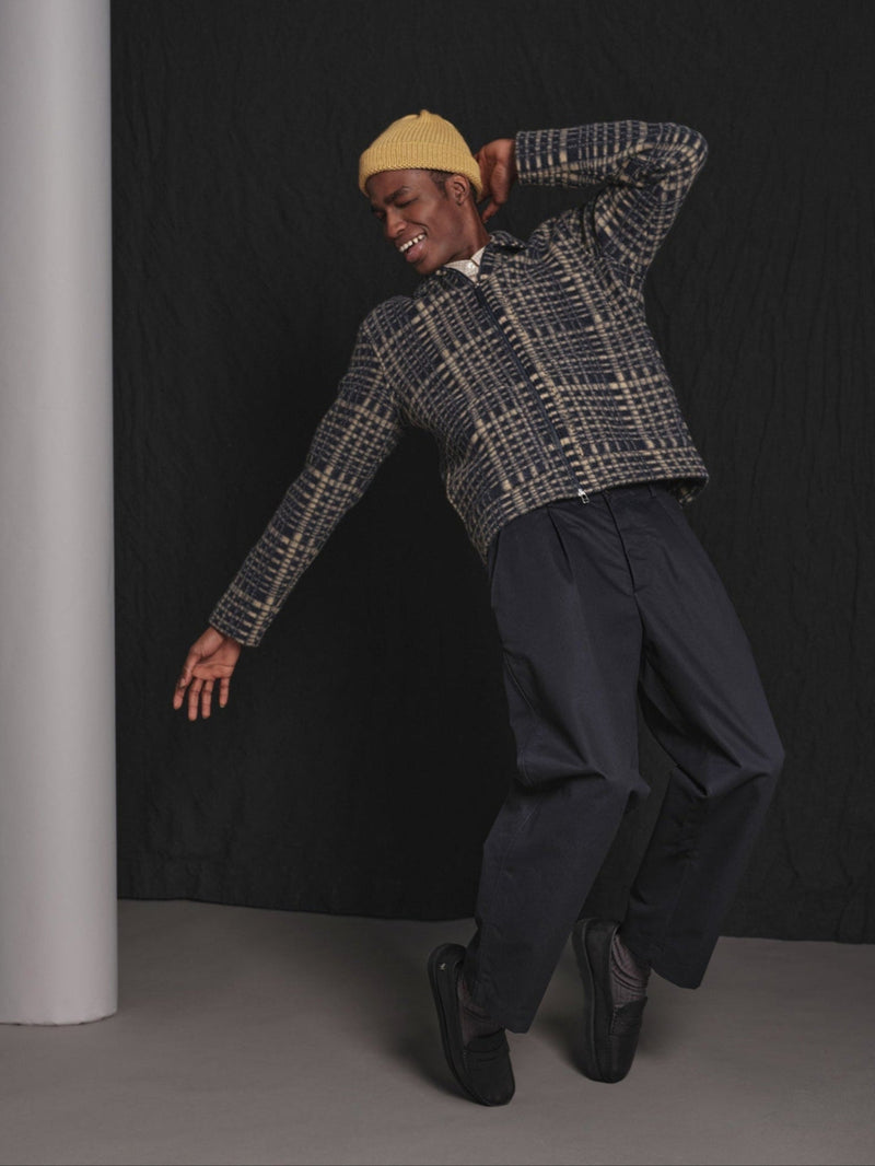 Person wearing a patterned jacket and dark pants leaning on tiptoes against a black background