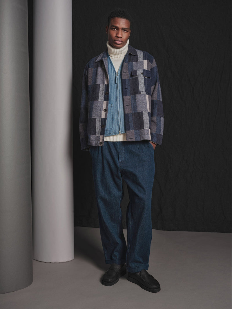 Man wearing a plaid shirt over a sweater with blue pants against a dark background