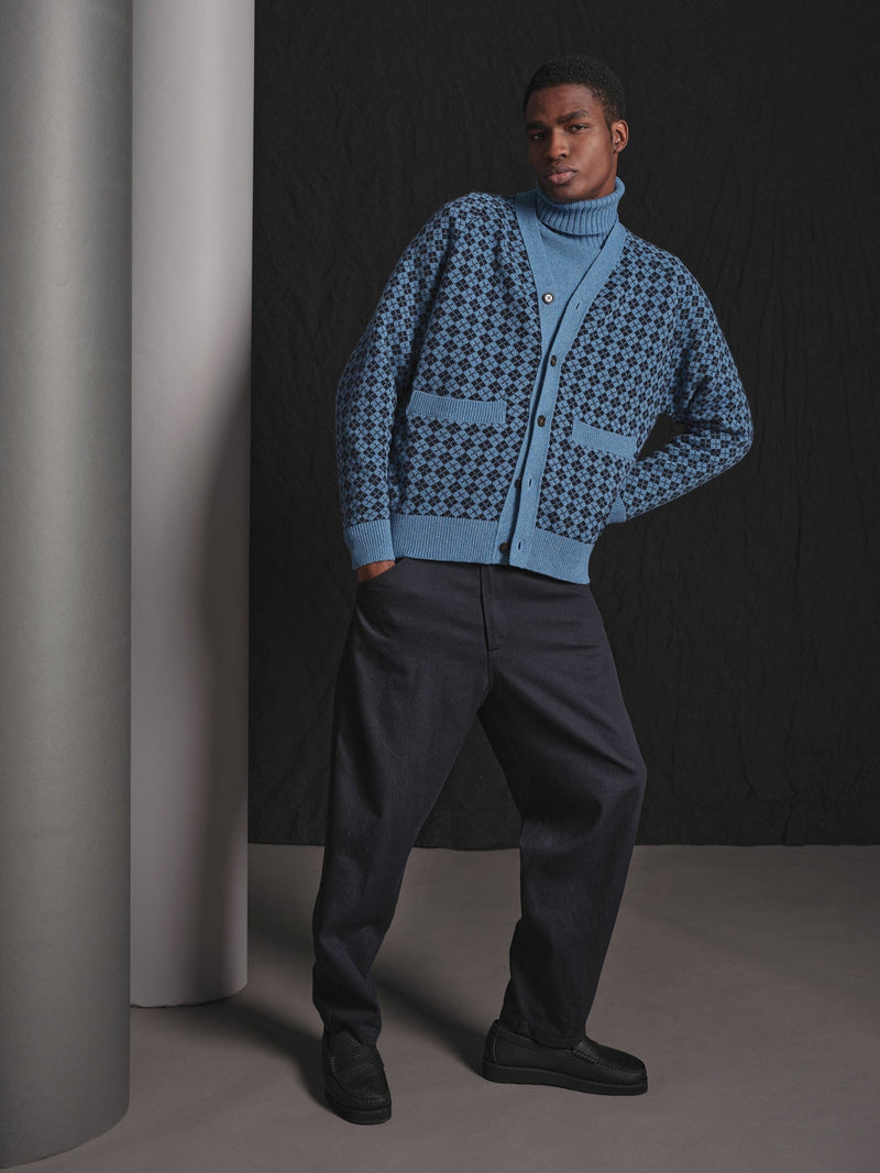 Man wearing a blue patterned cardigan and dark pants against a black background