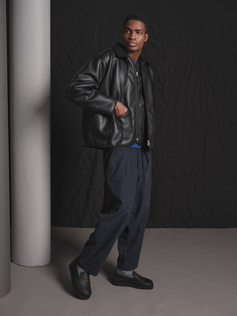 Man wearing a black faux leather jacket and dark pants against a black background