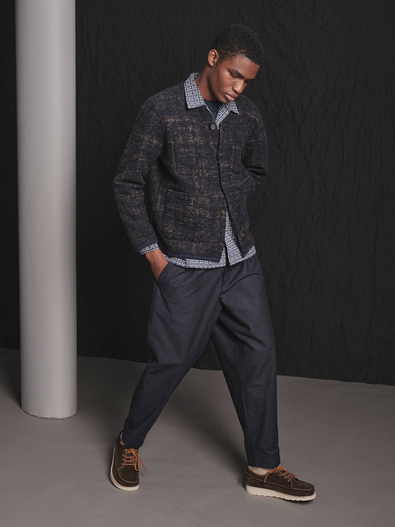 Man wearing a checkered shirt, gray cardigan, and dark pants against a black background