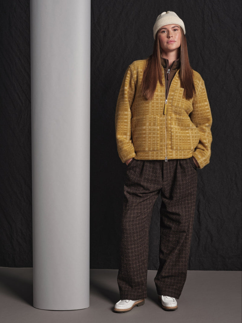 Person wearing a mustard yellow jacket and brown plaid pants standing against a dark background.