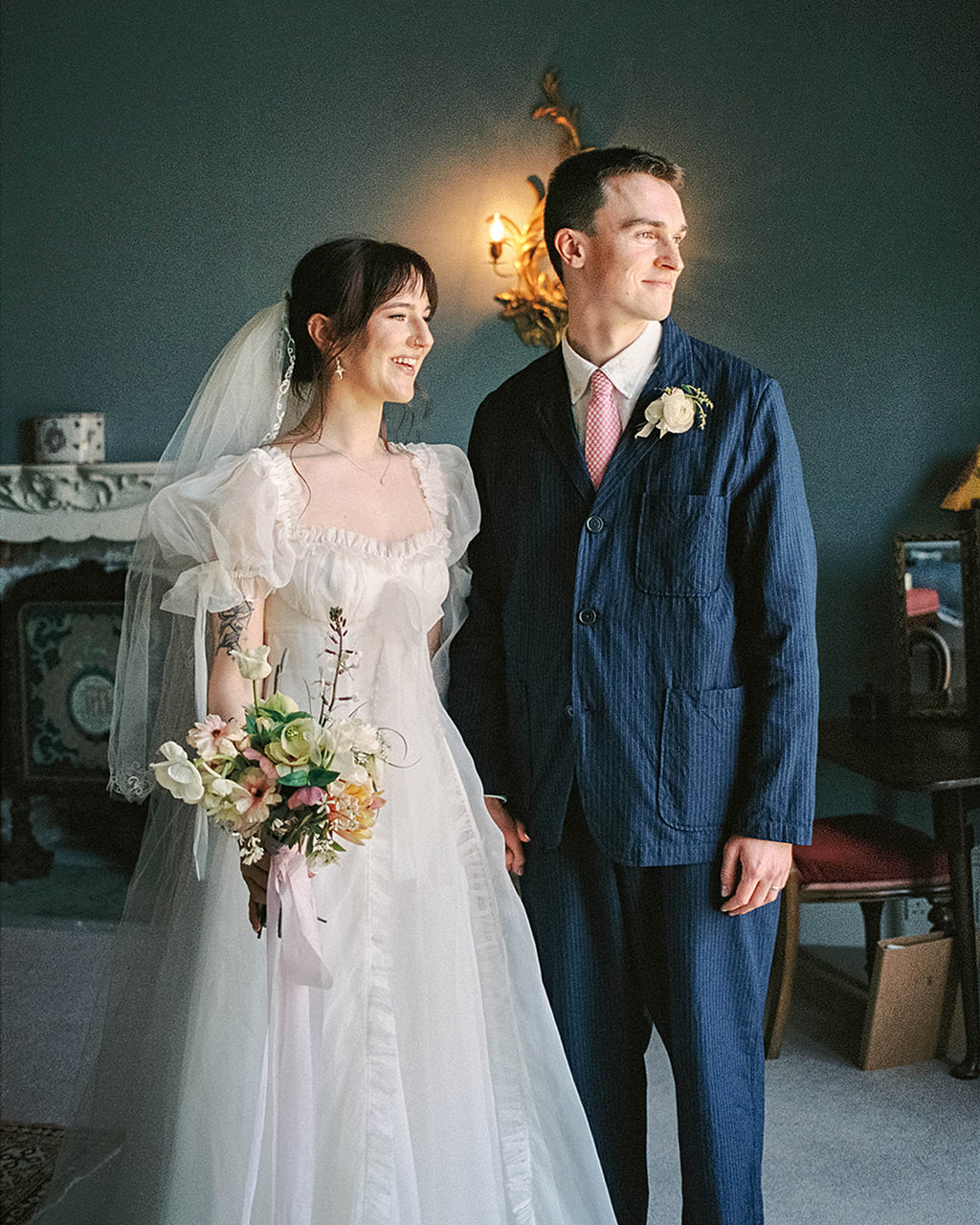 Couple in wedding attire standing in a room with a fireplace and decorative wall lights.
