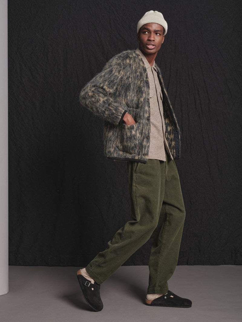 Man wearing a patterned jacket and green pants against a black background